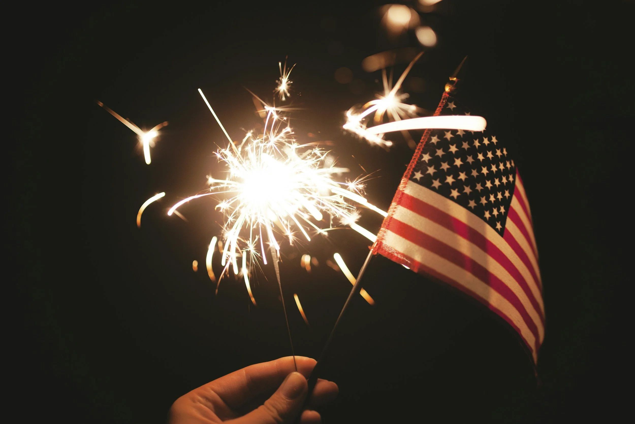 A photo of a hand holding both a small American flag and a lit sparkler at night.