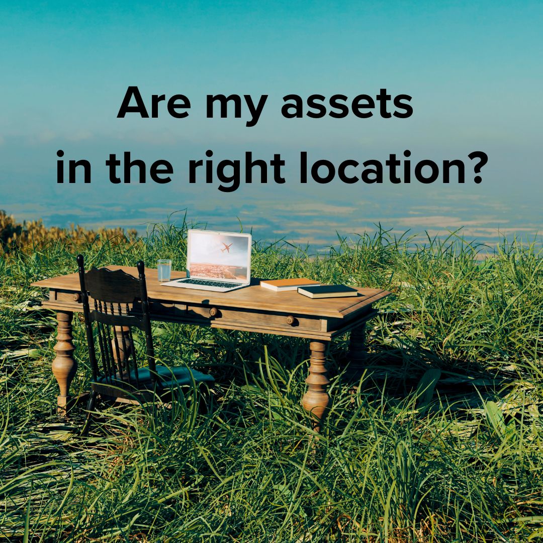 A desk, chair, computer, books, and a glass in tall grass on top of a hill overlooking a rural area. Black text at the top says: Are my assets in the right location?