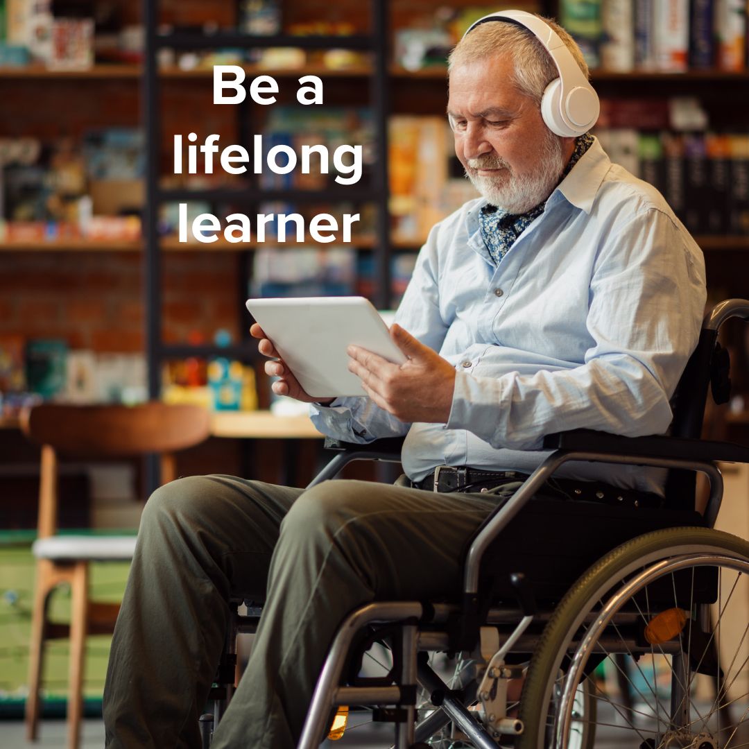 Man in a wheelchair in a library listening to headphones while watching a tablet. White text in the upper-left says: 'Be a lifelong learner'