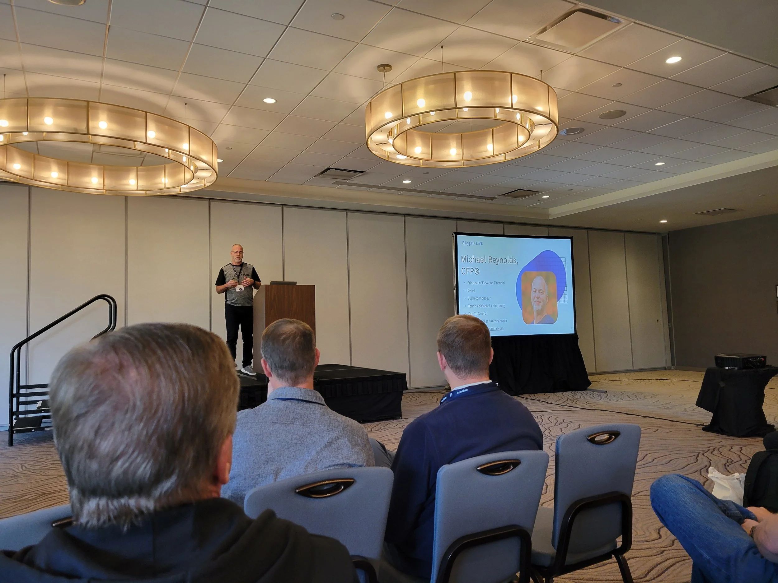 Photo of Michael Reynolds presenting to a crowd in the front of a large hotel conference room.