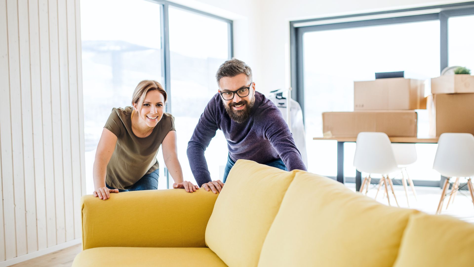 A yellow sofa being pushed across the room by a smiling heterosexual caucasian couple in their late 30s. Moving boxes rest on a dining table behind them.