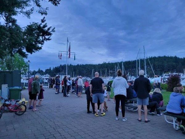 The Roche Harbor, Washington marina at dusk during the summer. A hill with pine trees are behind a row of sailboats and various flags. Many caucasian people sit or stand facing the flags.