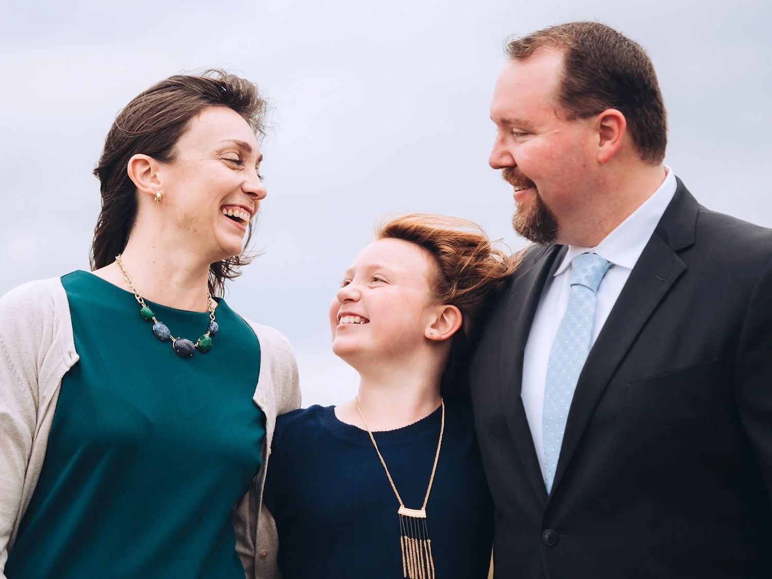 Three people standing, smiling. Kevin Estes on the right wearing a dark blue suit. Wife on the left wearing a dark green dress. Younger daughter in between wearing a dark blue dress. Light blue background.