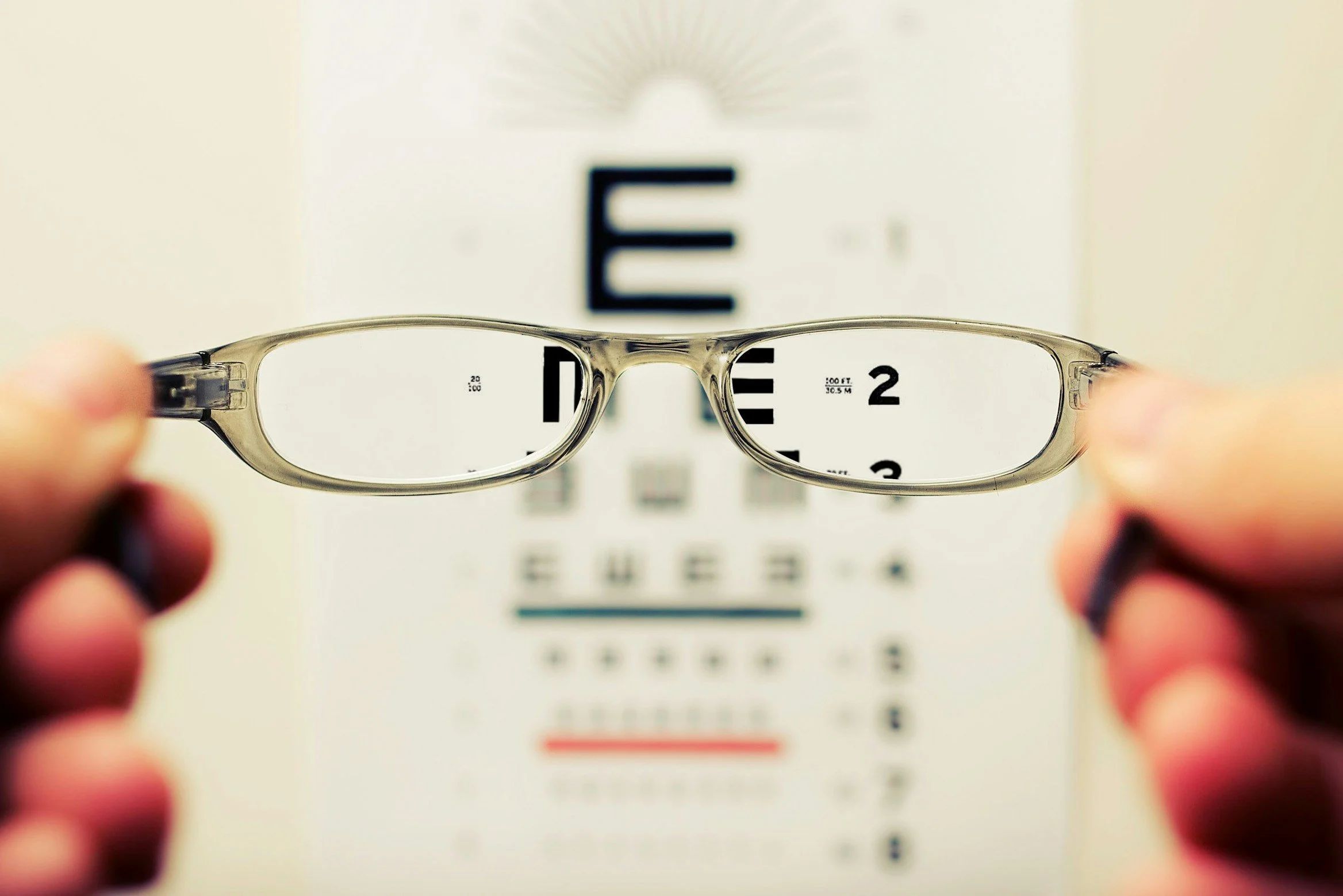 Photo of someone holding eyeglasses. The background is a blurry eye chart. However, the lenses show parts of the eye chart clearly.