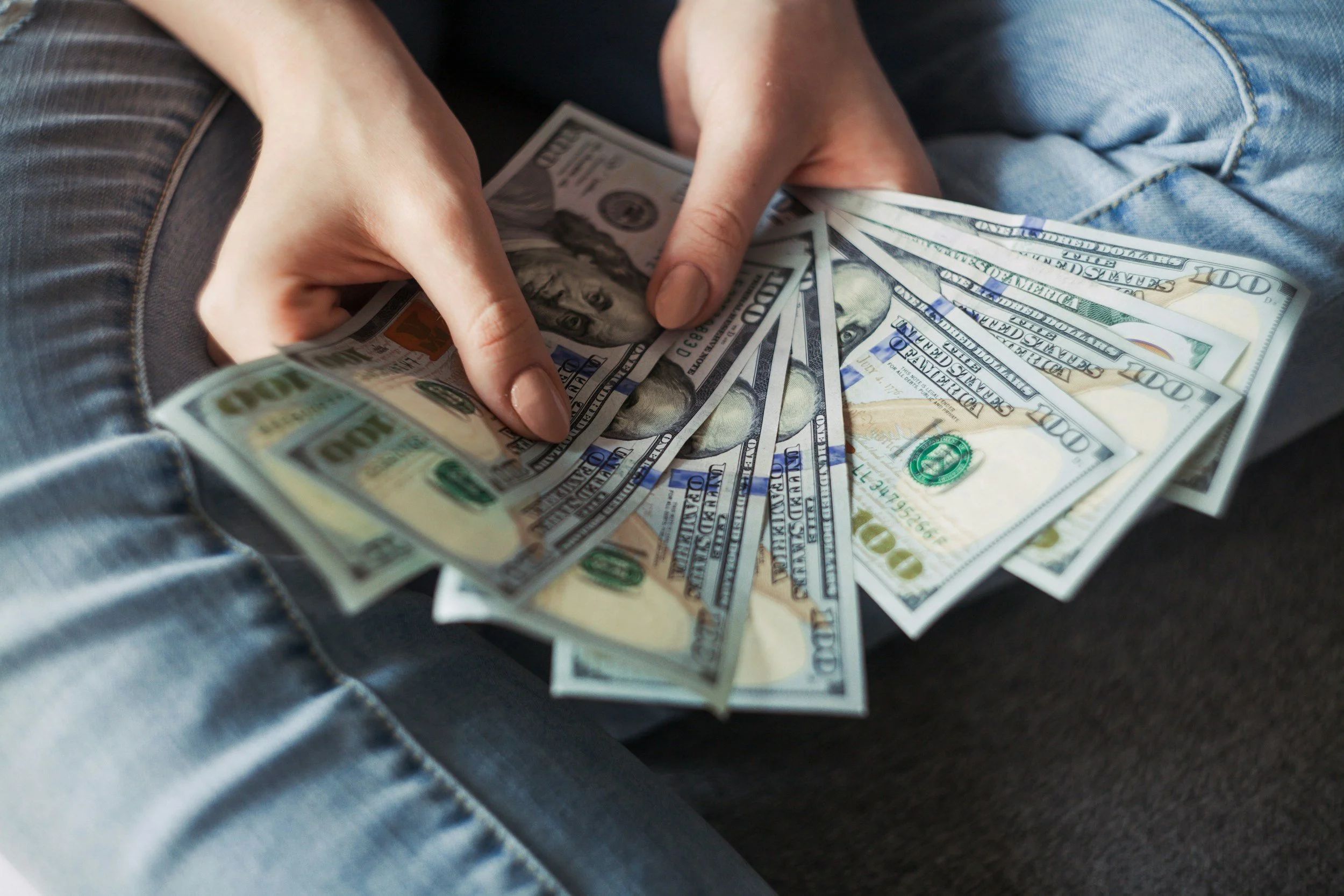 Photo of someone sitting in jeans and holding nine $100 bills fanned out in two hands.