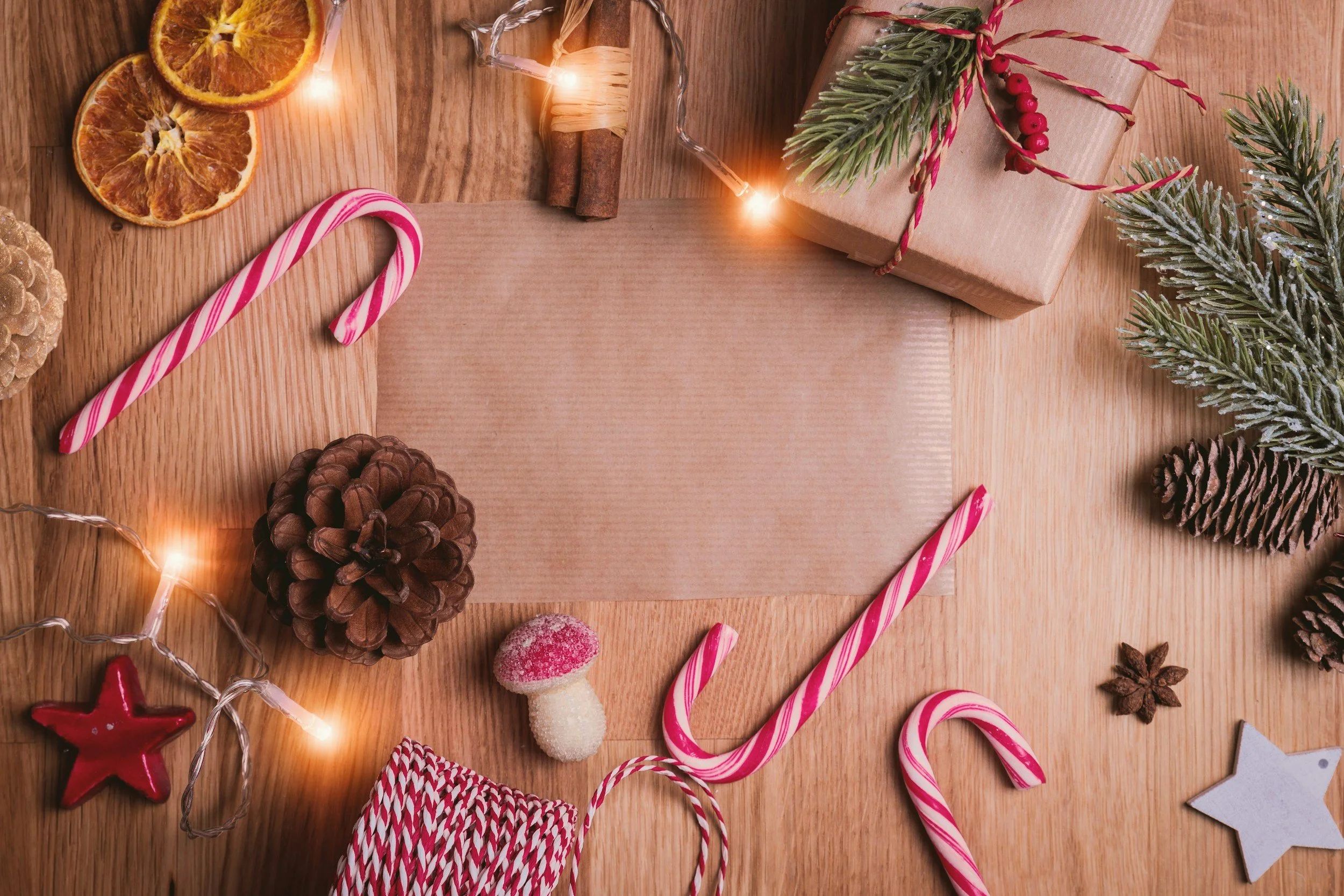 Photo on candy canes, white lights, pine cones, pine needles, decorative stars, twine, dried orange slices, star anise, and cinnamon on a wooden table.