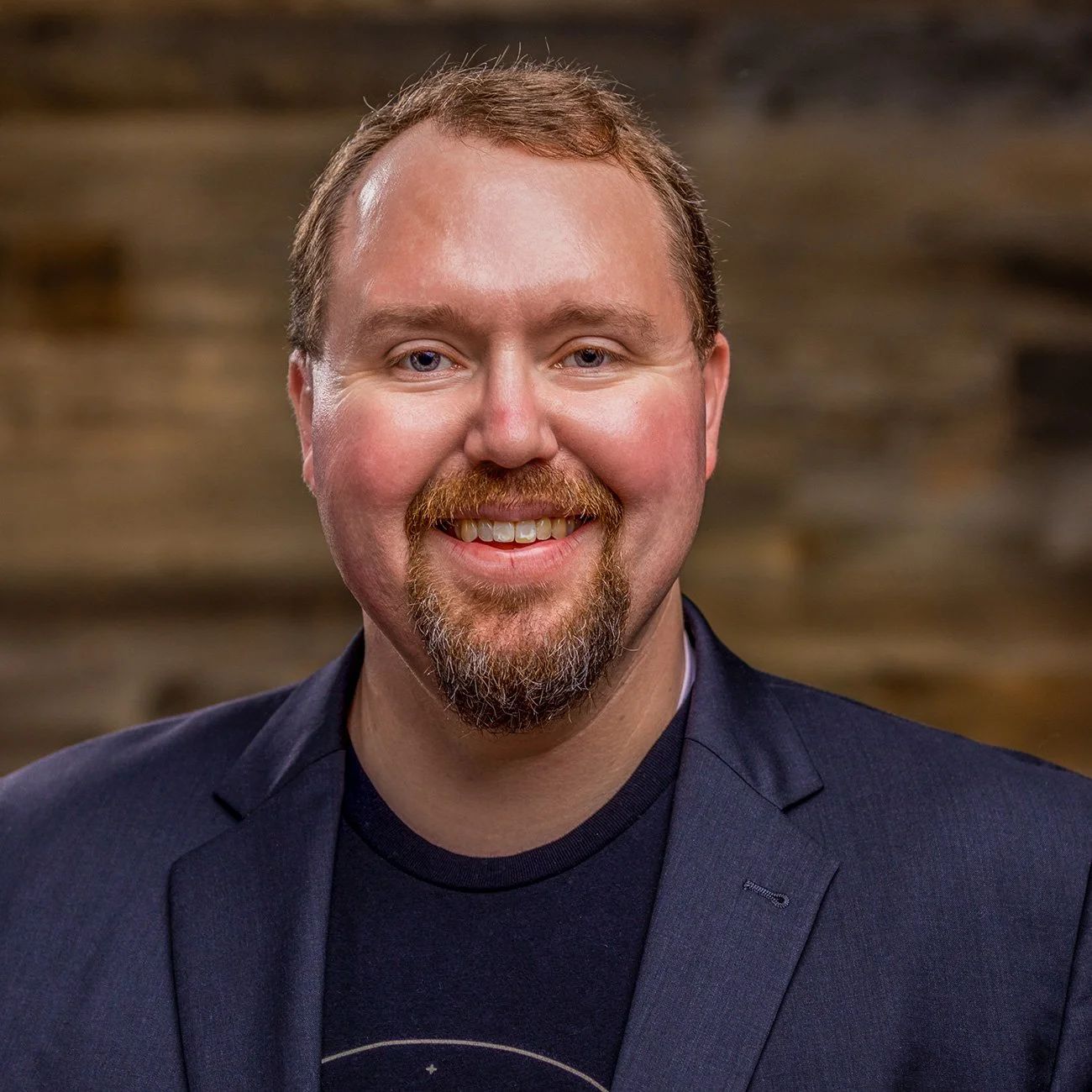 Photo of Kevin Estes smiling and wearing a dark suit with a black tee shirt. Behind him is an out of focus wooden background.