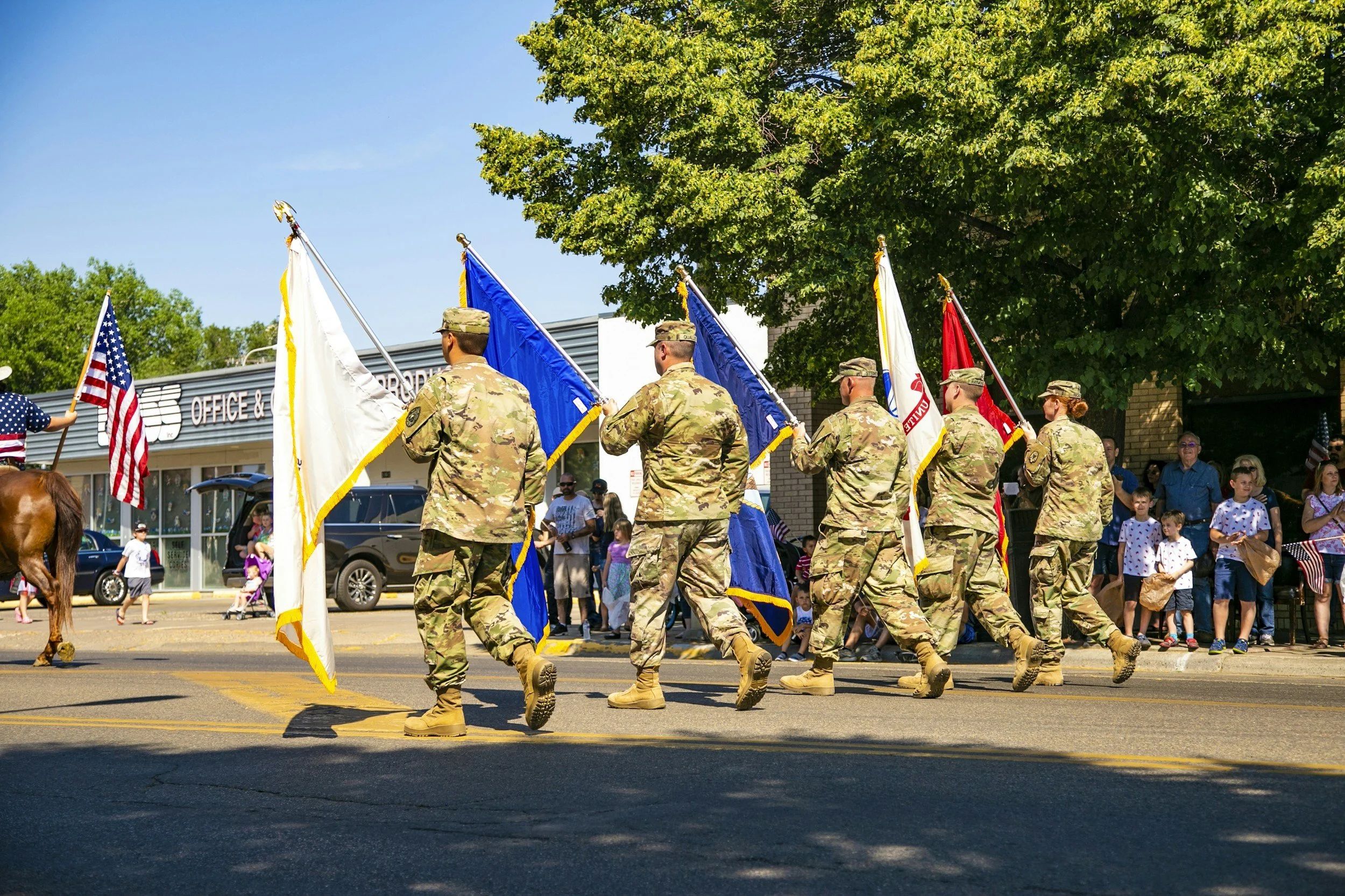 A photo of five soldiers carrying flags in a parade. A horse ridden by someone wearing an American flag shirt and carrying an American flag rides in front of them. Spectators stand in the shade of a large tree and watch.
