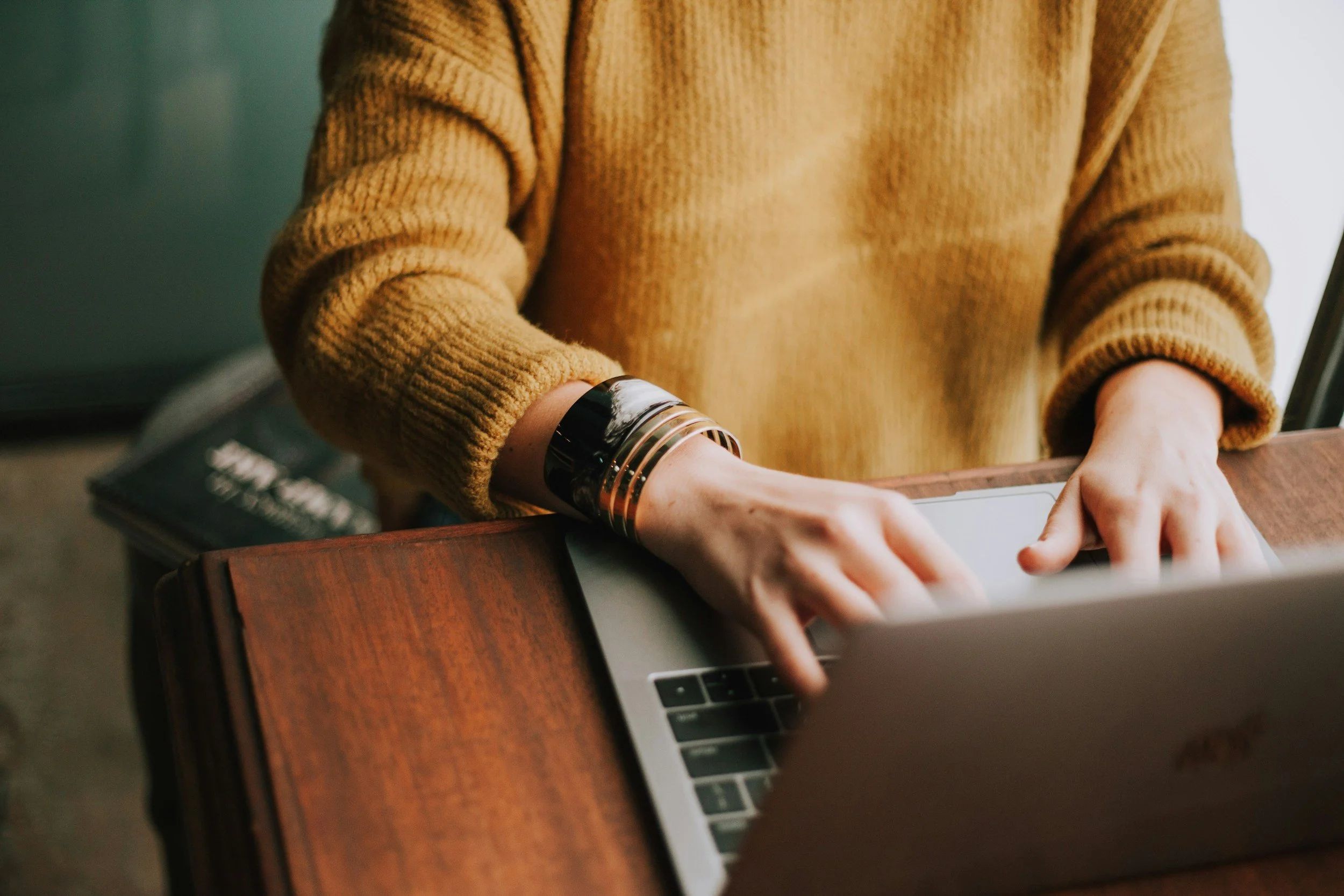 Photo of someone seated at a wooden desk and wearing a tan sweater who is typing on a laptop.